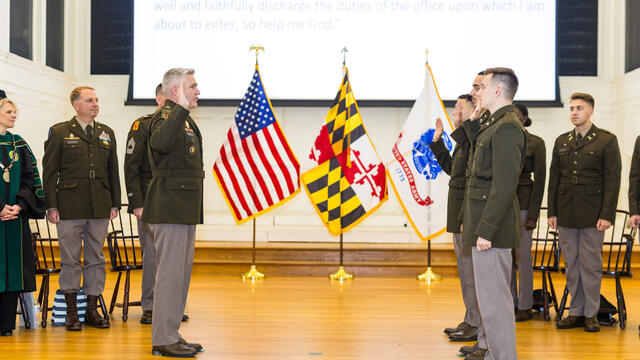 Photo of a stage with the U.S., Maryland, and Army flags and a row of cadets saluting an officer.