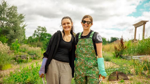 A student and professor pose outside in a garden while wearing gardening gloves.