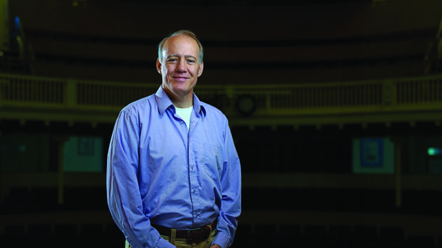 Professor Richard Sautter stands in a blue button up in the campus theatre.