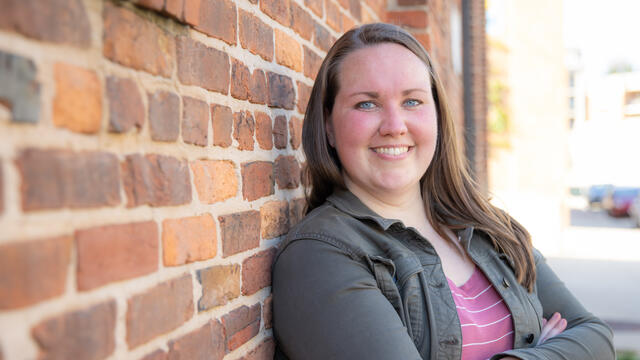 Photo of Robyn Forney standing in front of a brick wall, arms crossed, wearing a dark green button up and a pink shirt.