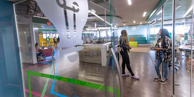 Two students talk at the end of a hallway with glass wall offices. A decal of a lightbulb is on one wall.